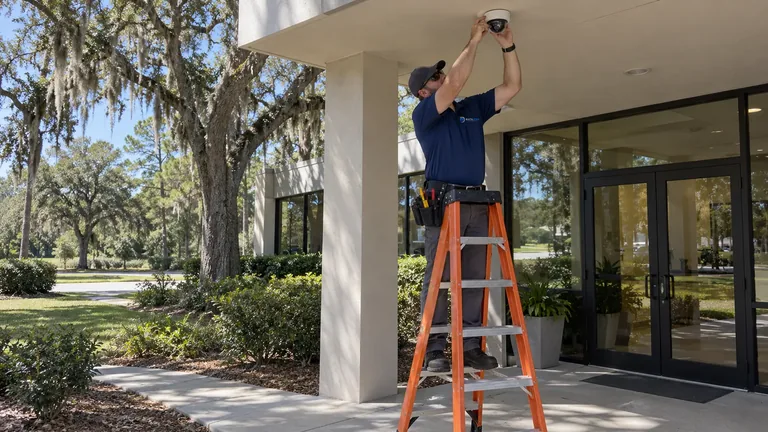Data Pro technician on an orange ladder installing a dome camera under the entrance canopy of a single-story commercial building shaded by live oaks