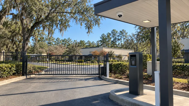Commercial vehicle gate lane with a slide gate, video intercom pedestal and a dome camera under the gate canopy among live oaks and pines