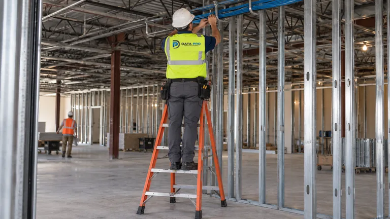 Data Pro technician in hard hat and vest on an orange ladder placing low-voltage cable into overhead J-hooks on a commercial jobsite